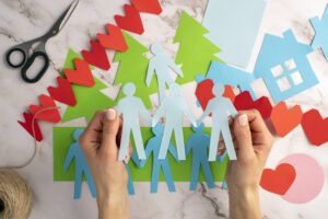 Schützende Hände umkreisen Holzfiguren in Menschenform, die im Kreis auf einem Holztisch stehen.